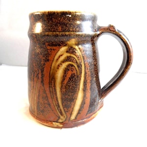 May include: A cylindrical ceramic mug featuring a dark brown and reddish-orange mottled glaze. The exterior surface displays a prominent, lighter-coloured arch-shaped pattern. It includes a sturdy, curved handle attached to the side of the vessel.