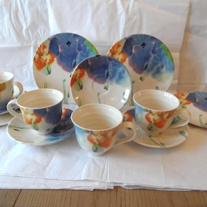 May include: A set of ceramic tea cups and saucers featuring a watercolour-style floral pattern with vibrant blue and orange petals on a white background. The collection includes multiple matching cups and saucers arranged together in a display.