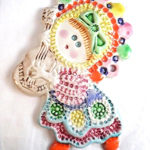 May include: A vibrant ceramic wall decoration depicting a doll-like figure. The figure features a round face with pink cheeks, a yellow hat, and a dress with diverse, colourful patterns. The figure is holding a musical instrument.