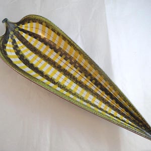 May include: A leaf-shaped ceramic dish featuring a vibrant pattern of alternating yellow and white stripes separated by dark brown bands. The edges of the dish are finished with a textured green speckled glaze, tapering to a point at one end.