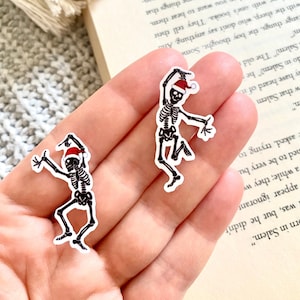 Peut inclure: Deux autocollants représentant des squelettes dansants coiffés de bonnets de Père Noël. Les squelettes sont en noir et blanc, avec des détails rouges sur les chapeaux. Les autocollants sont sur fond blanc, avec un livre et une surface neutre en arrière-plan.
