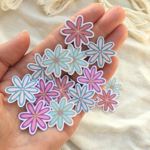 Peut inclure: Plusieurs autocollants en forme de fleurs aux couleurs pastel, rose, bleu et violet. Les autocollants sont tenus dans une main. Chaque fleur a un cercle central et un bord blanc. Le design est simple et joyeux.