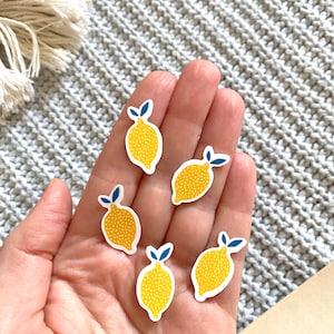 K&ouml;nnte beinhalten: Sechs gelbe Zitronen-Aufkleber mit blauen Bl&auml;ttern und wei&szlig;en Punkten auf einer Hand.