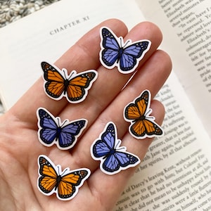 May include: Six butterfly stickers, three orange and three purple, on a hand holding a book. The book is open to a page with the text "CHAPTER XI".