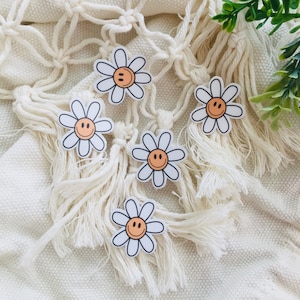 May include: Five white daisy stickers with orange smiley faces on a white crocheted background.