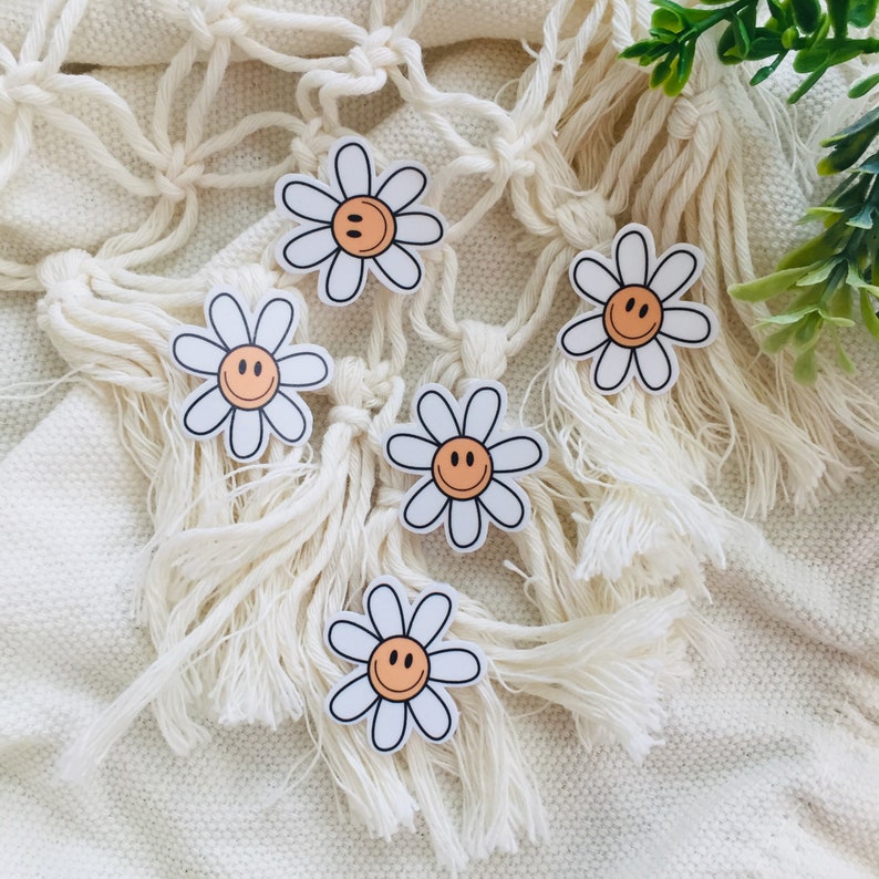 May include: Five white daisy stickers with orange smiley faces on a white crocheted background.