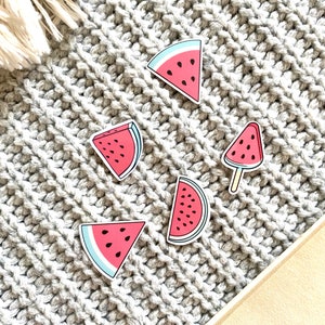 May include: Five watermelon stickers on a gray knitted surface. The stickers are in various shapes and sizes, including a slice, a wedge, and a popsicle.