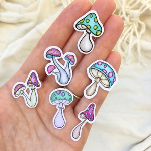 Peut inclure: Un ensemble de sept autocollants de champignons color&eacute;s sur fond blanc. Les champignons ont des chapeaux roses, violets, bleus et verts avec des points blancs. Les tiges sont blanches, bleu clair et vert clair.