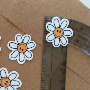 May include: Four white daisy stickers with a yellow smiley face in the center. The stickers are on a brown paper background.