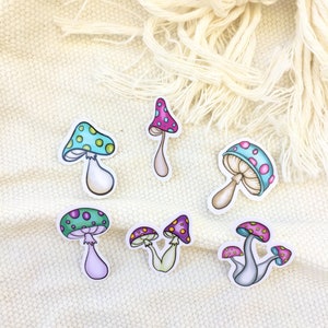 Peut inclure: Six autocollants en forme de champignon color&eacute;s avec des contours blancs. Les champignons ont des chapeaux de diff&eacute;rentes couleurs avec des pois blancs et des tiges. Les autocollants sont sur un fond de tissu blanc.