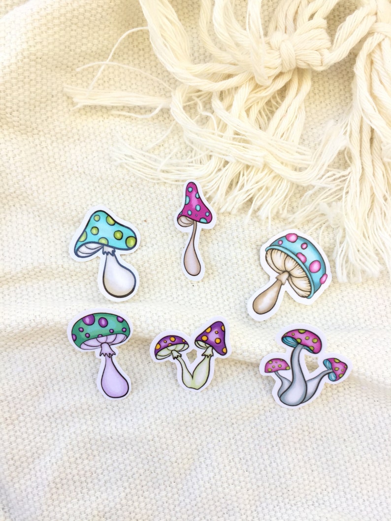 Peut inclure: Six autocollants en forme de champignon color&eacute;s avec des contours blancs. Les champignons ont des chapeaux de diff&eacute;rentes couleurs avec des pois blancs et des tiges. Les autocollants sont sur un fond de tissu blanc.