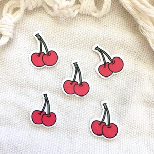 May include: Five red cherry stickers with green stems on a white fabric background.