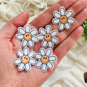 May include: Five white daisy stickers with a yellow smiley face in the center. The stickers are on a hand.