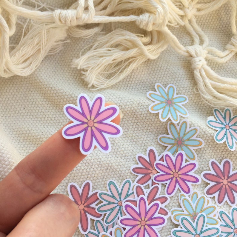 Peut inclure: Plusieurs autocollants de fleurs color&eacute;es dans des tons de rose, violet et bleu. Les autocollants ont un bord blanc et sont dispos&eacute;s sur une surface textur&eacute;e blanc cass&eacute;. Un autocollant est tenu par un doigt. Ces autocollants sont une fourniture artisanale.