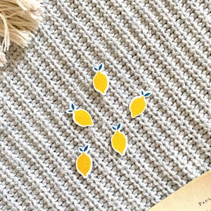 K&ouml;nnte beinhalten: F&uuml;nf gelbe Zitronenaufkleber mit gr&uuml;nen Bl&auml;ttern und wei&szlig;en Punkten auf einem grauen Strickhintergrund.