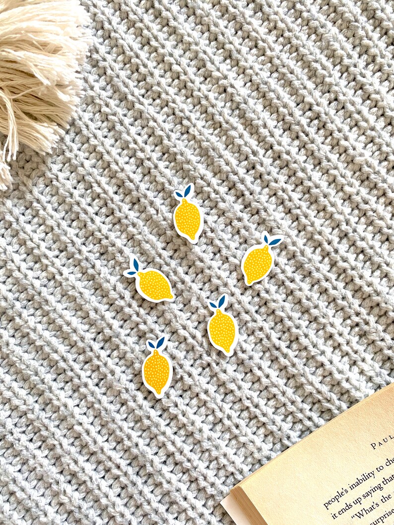 K&ouml;nnte beinhalten: F&uuml;nf gelbe Zitronenaufkleber mit gr&uuml;nen Bl&auml;ttern und wei&szlig;en Punkten auf einem grauen Strickhintergrund.