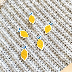 K&ouml;nnte beinhalten: F&uuml;nf gelbe Zitronenaufkleber mit blauen Bl&auml;ttern und wei&szlig;en Punkten auf einer grauen Strickoberfl&auml;che.