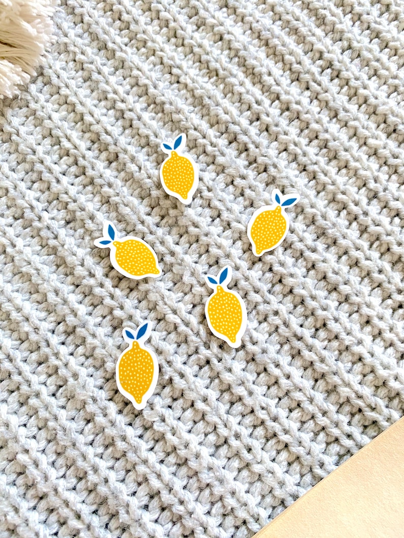 K&ouml;nnte beinhalten: F&uuml;nf gelbe Zitronenaufkleber mit blauen Bl&auml;ttern und wei&szlig;en Punkten auf einer grauen Strickoberfl&auml;che.
