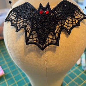 May include: A black lace bat with red gem eyes is pinned to a mannequin head. The bat has detailed wings with a spiderweb design. The head is covered in beige fabric, and the background is a green cutting mat.