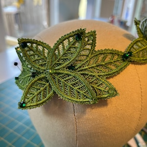 May include: A green lace leaf crown with dark green gemstones. The crown is made of delicate, open-weave lace and is adorned with small, round green gems. The leaves are arranged in a symmetrical pattern, creating a nature-inspired headpiece.