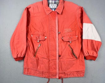 May include: A coral-colored jacket featuring a collar, long sleeves with red ribbed cuffs, and a white panel on the left sleeve. It includes two chest pockets with button flaps, two zippered waist pockets, a front button placket, and a drawstring hem.