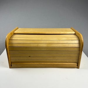 May include: A rectangular light brown wooden bread box featuring a curved roll-top lid constructed from horizontal slats. The storage container has solid side panels and a flat top surface, providing a secure enclosure for baked goods.