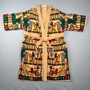 Puede incluir: Una bata de tela de rizo beige con un patrón de cuadrícula repetitivo de motivos de inspiración egipcia en rojo, amarillo, verde y negro. La bata incluye mangas largas, cuello de chal y un cinturón a juego, todo con textura acolchada.