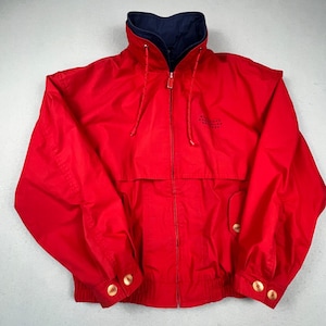 May include: A bright red zip-up jacket featuring a high collar with a dark blue interior lining. The jacket includes a full-length front zipper, long sleeves with buttoned cuffs, two front pockets with button closures, and thin drawstrings at the neck.