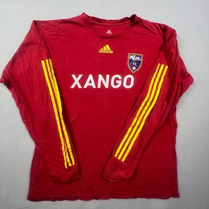 May include: A long-sleeve red athletic shirt featuring three yellow stripes down each sleeve. The front displays a yellow Adidas logo, the text XANGO in white, and a Real Salt Lake team crest with a crown, the letter R, and a flower emblem.