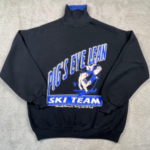 May include: Black sweatshirt with a blue turtleneck collar. The front features the text "PIG'S EYE LEAN SKI TEAM" in white and blue, along with a cartoon pig skiing. The sweatshirt has long sleeves.