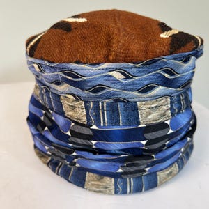 May include: A cylindrical fabric hat featuring a brown textured top with dark brown and white accents. The sides are constructed from multiple horizontal bands of blue, grey, and white patterned fabric, showcasing various geometric and wave designs.