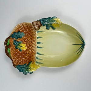 May include: A ceramic serving dish shaped like an acorn with a textured brown cap and a smooth light yellow base. The dish features sculpted green and yellow oak leaves and small acorns along the edge, with painted green lines on the interior.