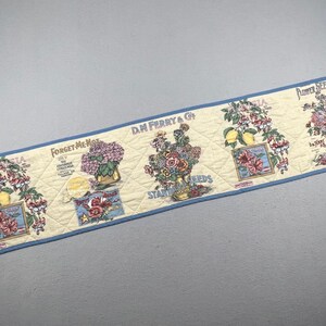 May include: A quilted table runner with a cream background and blue trim, featuring repeating botanical illustrations of flowers and lemons. The fabric displays vintage-style seed packet graphics with text including D.M. Ferry & Co. and Forget-Me-Not.