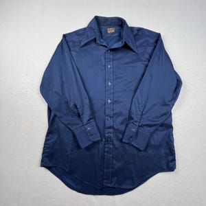 May include: A solid navy blue long-sleeve button-down shirt featuring a pointed collar, a single chest pocket on the left side, and matching navy blue buttons down the front placket and on the barrel cuffs. The shirt has a standard hemline.