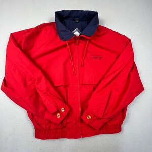 May include: A bright red windbreaker jacket featuring a navy blue fold-down collar, a full-length front zipper, and long sleeves with buttoned cuffs. The jacket includes a ribbed hem, a drawstring at the neck, and small black text on the chest.