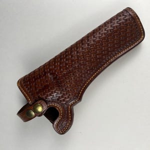 Pode incluir: Um coldre de couro castanho com um padrão detalhado de entrelaçamento gravado na superfície. O coldre inclui costuras de cor clara ao longo das bordas e uma pequena alça fixada na base com dois botões de pressão de metal na cor latão.