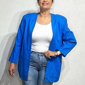 Könnte beinhalten: Ein leuchtend blauer Blazer mit langen Ärmeln und lockerer Passform. Der Blazer wird offen getragen und zeigt ein weißes Rippen-Tanktop und blaue Jeans. Das Outfit wird durch eine Uhr und Armbänder ergänzt.