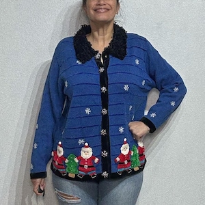 Puede incluir: Un cárdigan navideño azul con cuello y ribetes negros. El cárdigan presenta diseños de copos de nieve y Papá Noel. El cárdigan tiene mangas largas y se abrocha por delante.