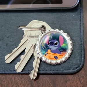 May include: A silver key chain with a blue Stitch character from Lilo & Stitch on a bottle cap charm. The charm has a colorful background with a tropical scene.