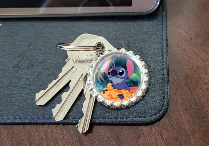May include: A silver key chain with a blue Stitch character from Lilo & Stitch on a bottle cap charm. The charm has a colorful background with a tropical scene.