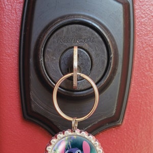 May include: A silver key chain with a blue Stitch character from the Disney movie Lilo & Stitch on a bottle cap charm. The keychain is attached to a keyhole on a black door handle with the word "Kwikset" on it.