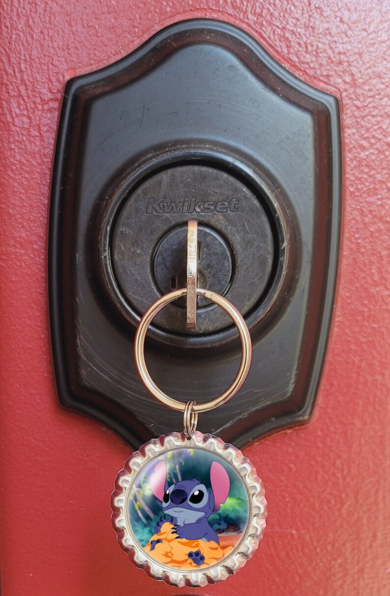 May include: A silver key chain with a blue Stitch character from the Disney movie Lilo & Stitch on a bottle cap charm. The keychain is attached to a keyhole on a black door handle with the word "Kwikset" on it.