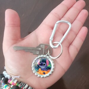 May include: A silver key chain with a blue Stitch character on a bottle cap charm. The keychain also has a silver carabiner clip.