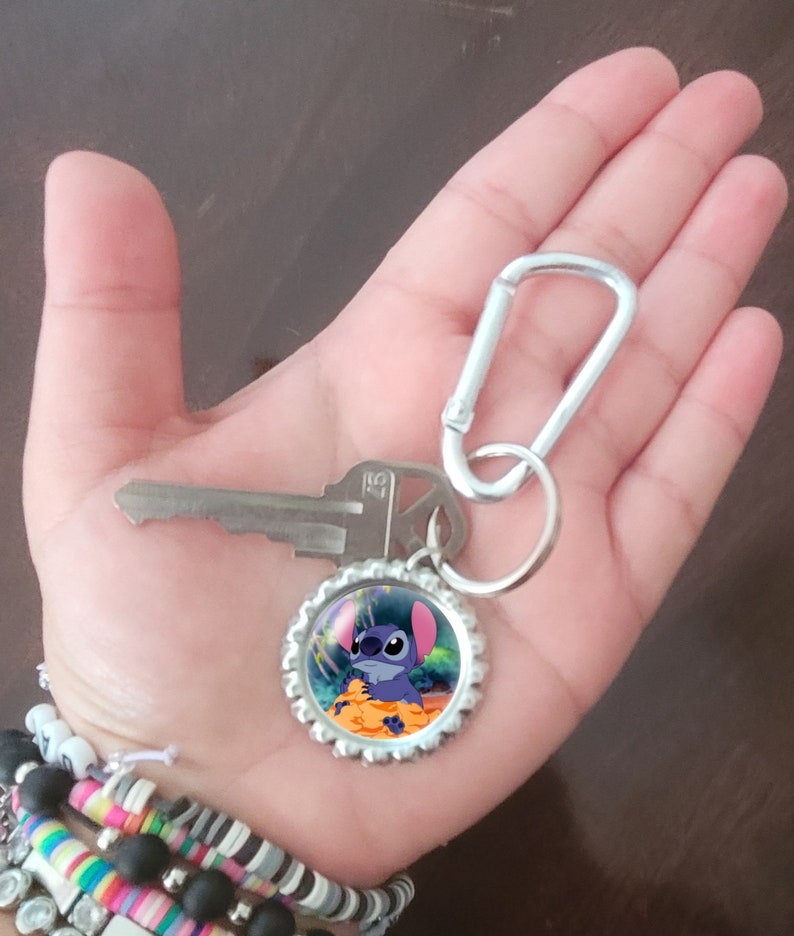 May include: A silver key chain with a blue Stitch character on a bottle cap charm. The keychain also has a silver carabiner clip.