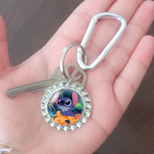 May include: A silver key chain with a blue Stitch character on a bottle cap charm. The charm has a colorful background with a green, purple, and orange design.