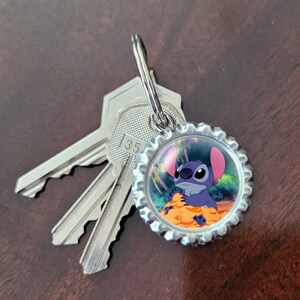 May include: A silver key chain with a blue Stitch character from the Disney movie Lilo & Stitch on a bottle cap charm. The charm has a colorful background with palm trees and a beach scene.