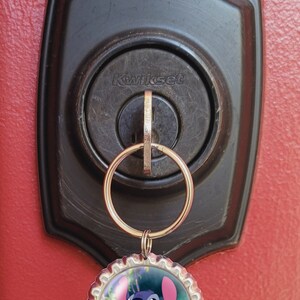May include: A silver key chain with a blue Stitch character from the Disney movie Lilo & Stitch on a bottle cap charm. The keychain is attached to a keyhole on a black door handle with the word "Kwikset" on it.