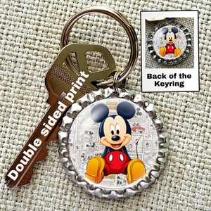 May include: A silver bottle cap keychain with a double-sided print of Mickey Mouse. One side features Mickey Mouse sitting with a comic book background. The other side features Mickey Mouse in a simple pose. The keychain is attached to a gold key ring.
