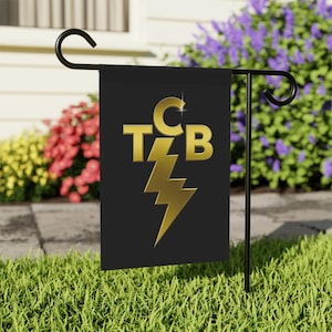 Puede incluir: Una bandera de jardín negra con las letras doradas "TCB" y un diseño de rayo dorado. La bandera se muestra en un soporte de metal negro en un jardín cubierto de hierba, con flores coloridas en el fondo.