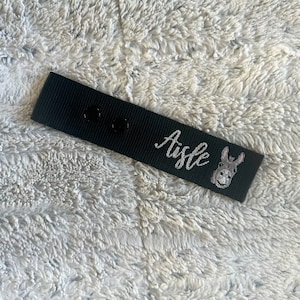 May include: Black fabric tag with the word "Aisle" in cursive script and a small donkey illustration. Two black snaps are visible on the tag. The tag is laying on a fluffy, white surface.